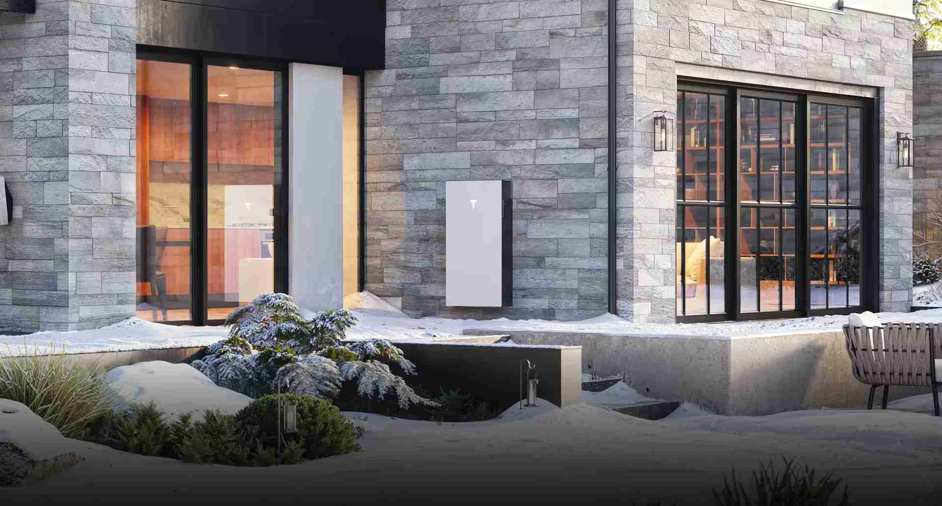 A modern, architect-designed house with a white Tesla Powerwall 3 battery unit mounted on its grey stone exterior wall during a snowy winter evening.