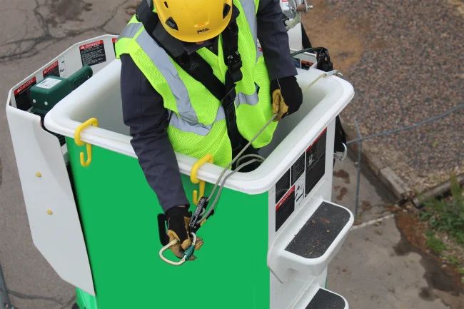 Close-up of a fall protection safety harness carabiner attached to an anchor point on a boom truck bucket against a clear sky